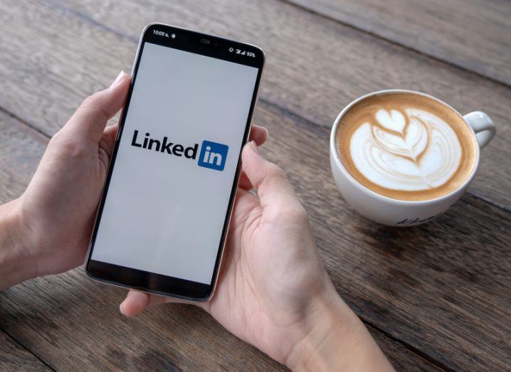Person's hands holding a phone with LinkedIn open on it and a cappuccino beside them.