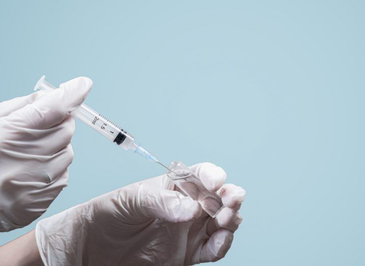 Hands in gloves holding a vaccine needle with a vial of clear liquid. Light blue background.