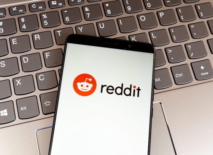 A mobile phone with the Reddit logo on the screen, resting on a laptop keyboard.