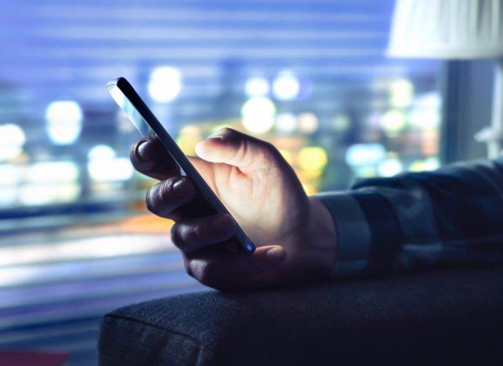 Person's hand using a mobile phone, resting on an armchair with a lamp and window in the background.