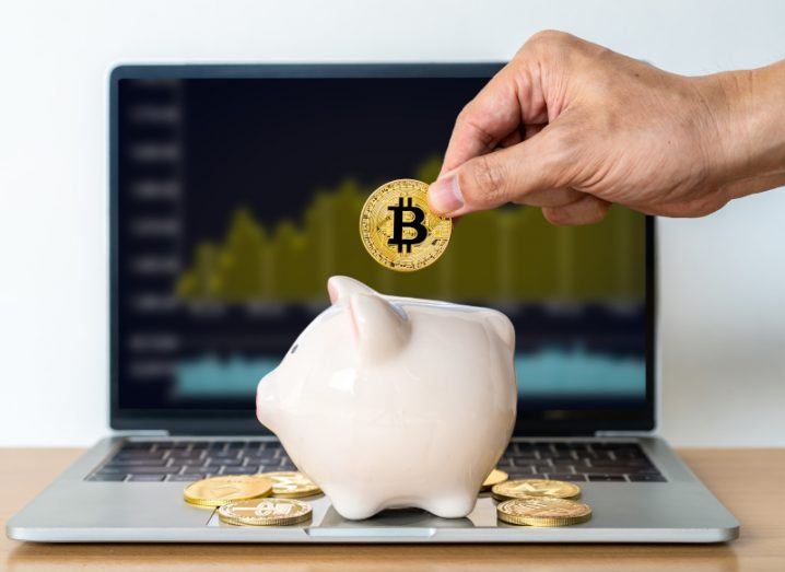 A hand holding a bitcoin, putting it into a piggy bank that is resting on a laptop with other coins around it.