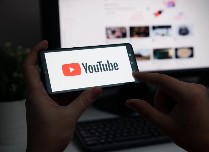 Two hands holding a phone with the YouTube logo on the screen, with a computer screen in the background showing YouTube videos.