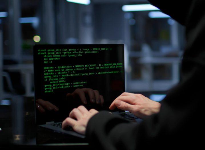A person wearing a black hoodie typing code on a laptop in an office environment.