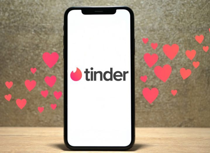 Tinder logo on a smartphone standing up on a wooden platform with orange hearts surrounding the phone.