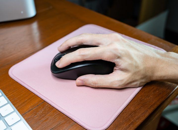 A hand on a mouse, resting on a pink mousepad on a wooden table.