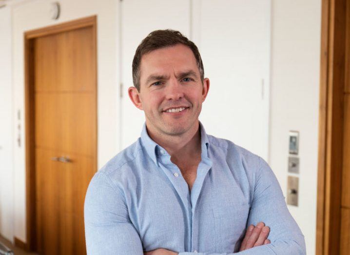 A man in a blue shirt with his arms crossed, standing with a cream wall and a brown door in the background. He is Conor O'Loughlin, CEO of Glofox.