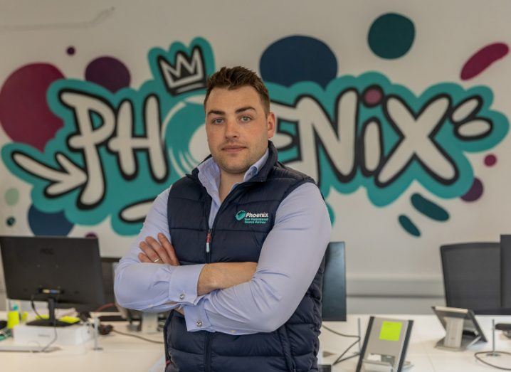 A man in a branded Phoenix recruitment gilet stands with his arms folded in front of a desk. The wall behind him has a graffiti mural of the word Phoenix.