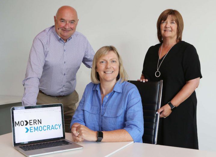 A woman seated with a man and a woman both standing behind her. They are gathered around a laptop with the Modern Democracy logo on it.