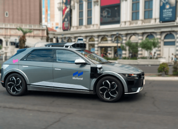 A grey electric vehicle with the Motional and Lyfy logos on the side, with buildings in the background.. It is an autonomous vehicle.