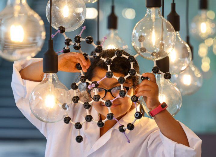 A young girl in a white lab coat holds up a model of molecules.