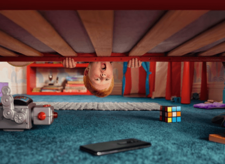A child looking under his bed and spotting a smartphone, with a toy robot and a Rubik's Cube.