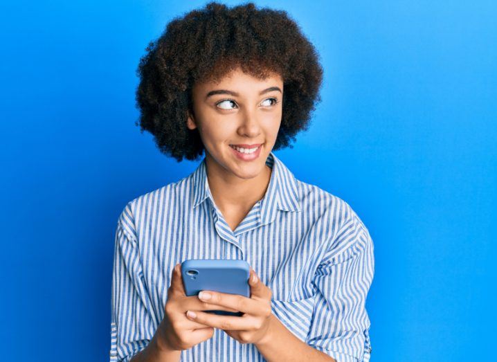 Young woman holding a phone in her hands and looking away in a blue background. The image symbolises users leaving groups on WhatsApp.