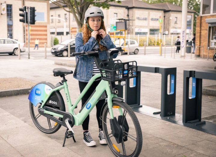 Woman wearing helmet is about to mount one of the ESB e-bikes in an outdoor area.
