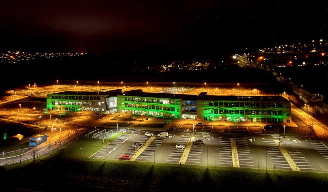 A night shot of the TCS Global Delivery Center in Letterkenny.  A vast building with many lights in the surrounding darkness.  The lights of other buildings can be seen in the distance.