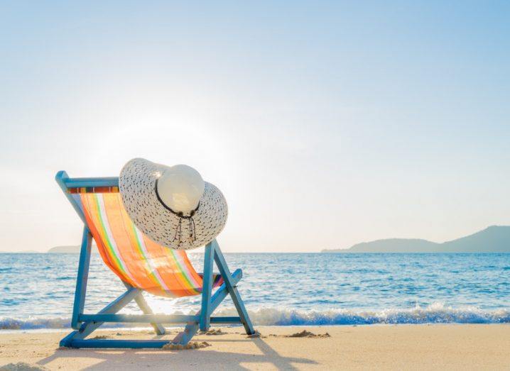 A deck chair at a sunny beach with a hat hanging from its side.