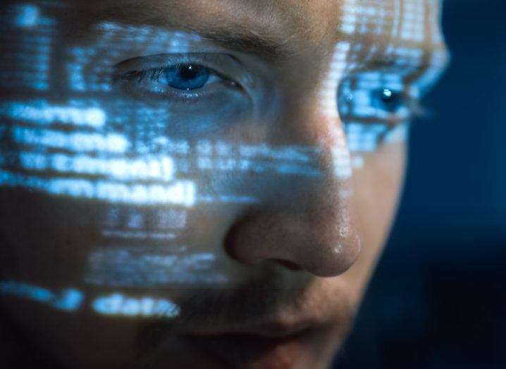 Man with blue eyes stares into a computer screen with reflections of code on his face.