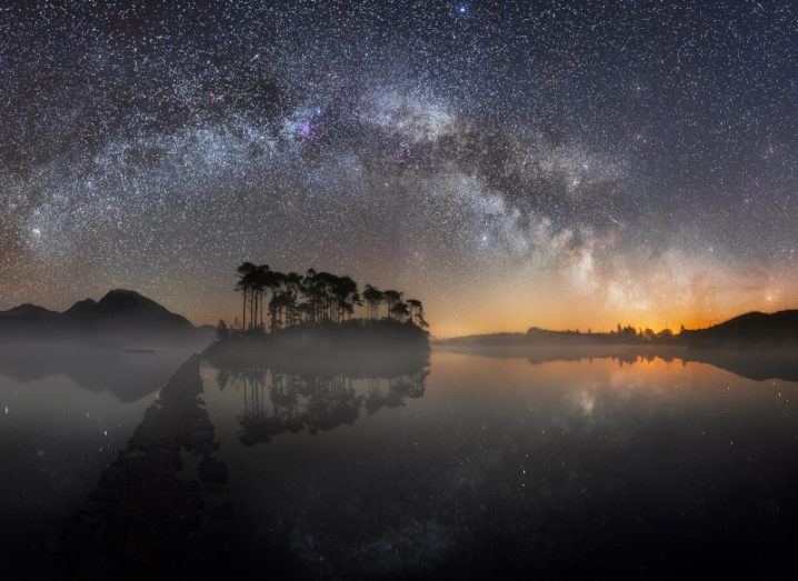 The Milky Way as seen from Pine Island in Connemara, Galway. A water body reflects the sky with trees visible in the distance.