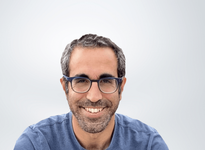 A man in a blue T-shirt smiles at the camera against a light grey background.