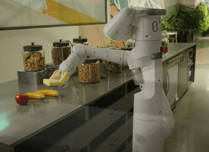 A robot picking up a sponge from an office kitchen counter, with an apple and bananas next to it. It is an Everyday Robot made by Alphabet.