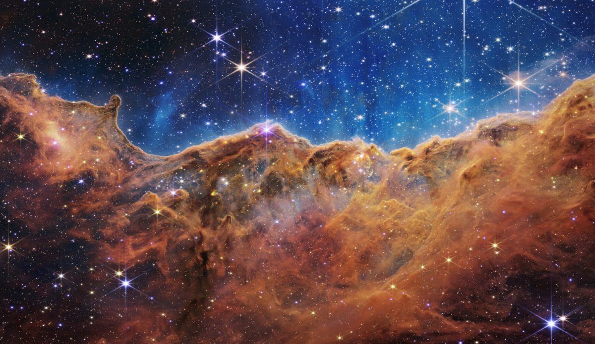 A mass of orange gas that ends as cliffs in the middle of the image and more stars above.  There are masses of stars visible in the gas.