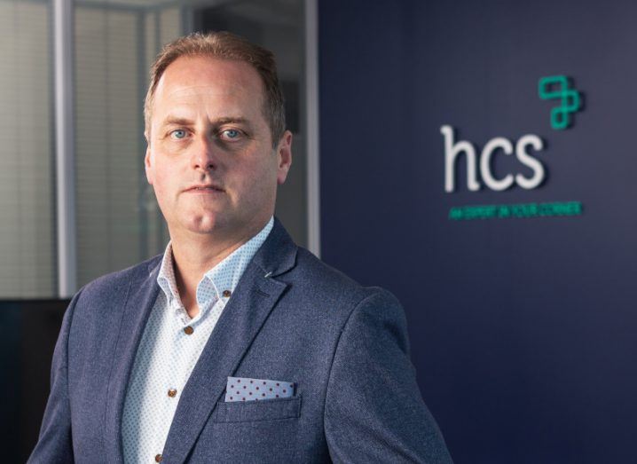Neil Phelan stands in a suit in front of a wall that has the HCS logo on it.
