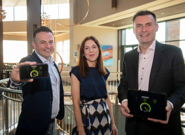 Two men and a woman who work for Siro, in the interior of an office. The two men are holding a smartphone and tablet that show internet connection speeds.