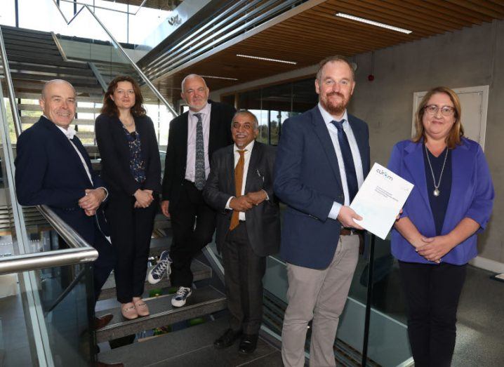 Four men and two women standing by stairs, with a man in front holding a white paper written by Cúram, the Science Foundation Ireland research centre.