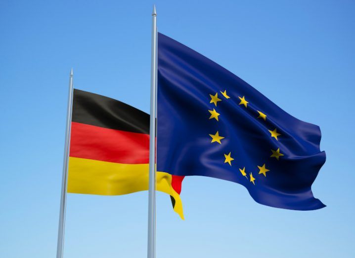 An EU flag waving in front of a German flag, with a clear blue sky background.