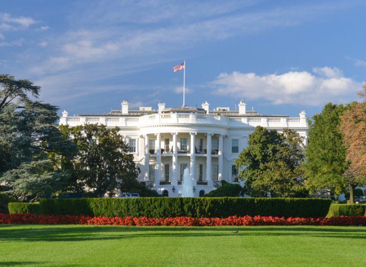 The White House with a US flag on top, with a clear sky in the background and a green garden with hedges and trees in front of it.
