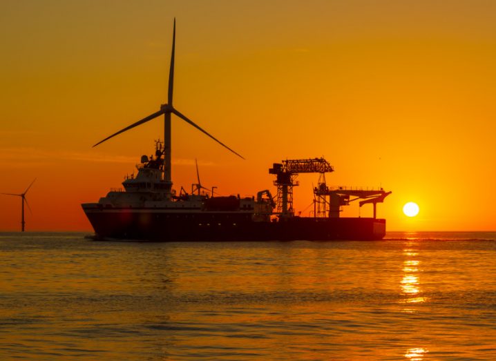 An offshore wind turbine with a boat in front of it, on the sea with the sun setting in the background.