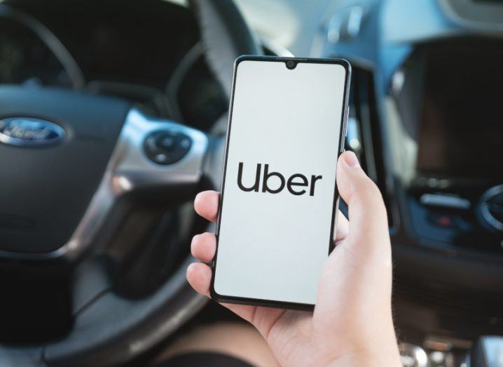 A person holding a smartphone with the Uber logo on the screen, inside a car with a steering wheel behind the phone.