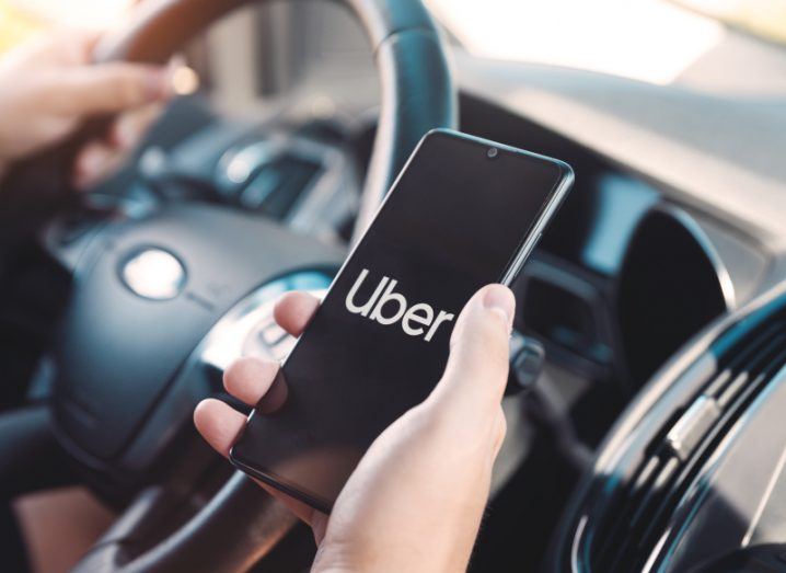 A person holding a mobile phone with the Uber logo on the screen, with the interior of a car and a steering wheel in the background.