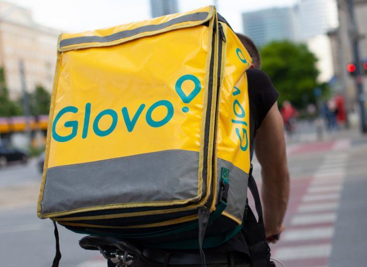 A person on a bike wearing a large bag with the Glovo logo on it. A city road and street is in the background.