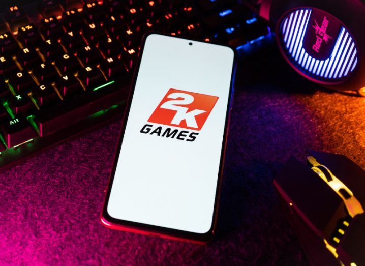 2K Games logo on a smartphone screen. The phone is resting on a table next to a keyboard and mouse.