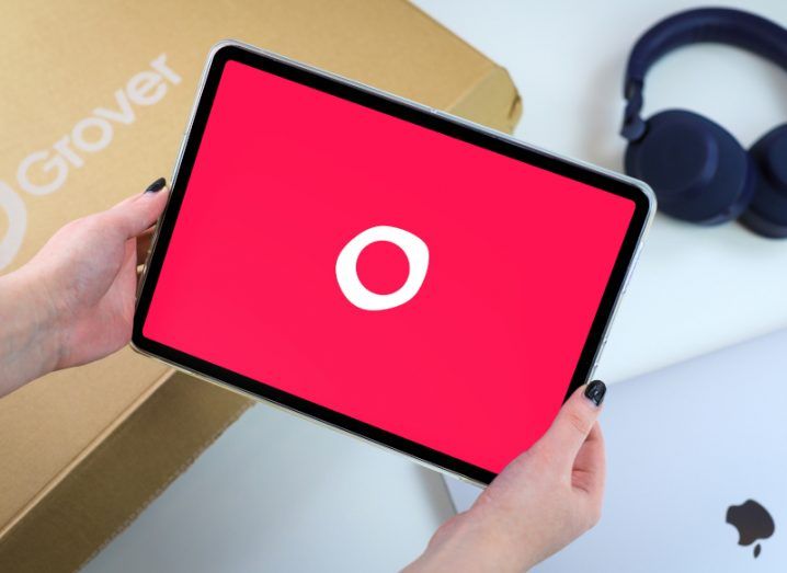 An iPad tablet with the Grover logo on the screen, held in a person's hands with a box and headphones laying on a white background.