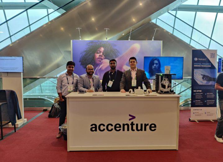 Four men standing behind a table with the Accenture logo on it, with a VR headset on the table and billboards and windows in the background.