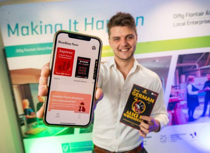 Evan McGloughlin holds up a phone displaying the Weeve Languages app, while holding a Weeve paperback of Alice in Wonderland for German language learners in his other hand.