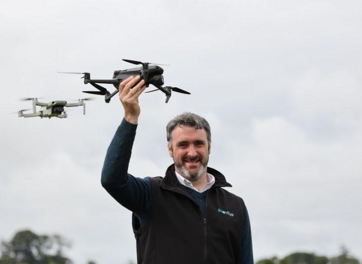 A man holing a drone above his head in one hand, with another drone flying in the background and a grey cloudy sky in the distance. He is Jerome O'Connell, CEO and co-founder of ProvEye.