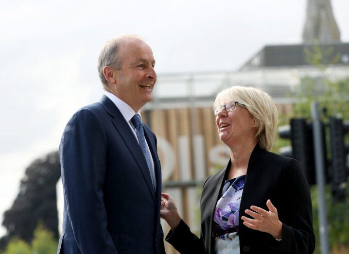 A man and a woman standing and laughing next to each other with a building in the background. They are Micheál Martin and Eeva Leinonen at Maynooth University.