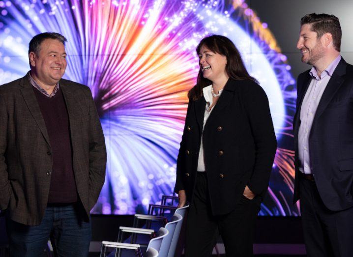Two men and a woman smile at each other in front of a large screen displaying a colourful strobe-like design.