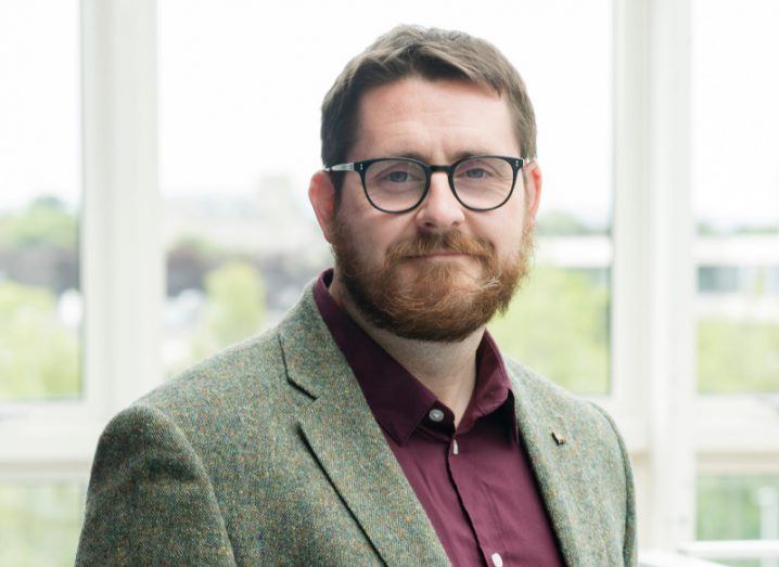 Headshot of Dr Steve Campbell wearing a moss-coloured tweed blazer and maroon shirt, standing in front of a large window.