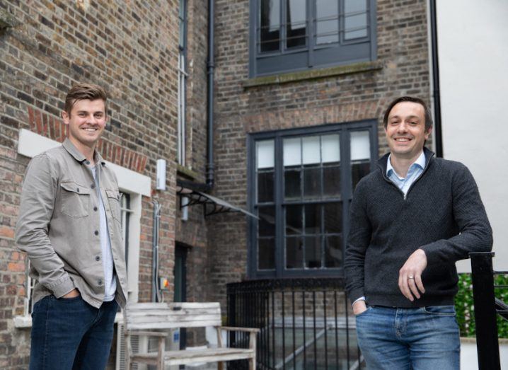 Two men standing in front of a brick building with windows and a gate in the background. They are Wayflyer co-founders Jack Pierse and Aidan Corbett.
