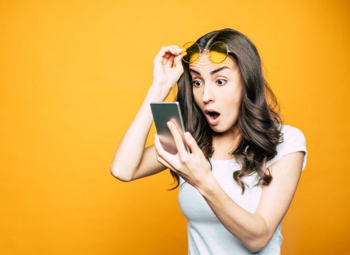 Woman raises her glasses and looks at a smartphone screen with shock.