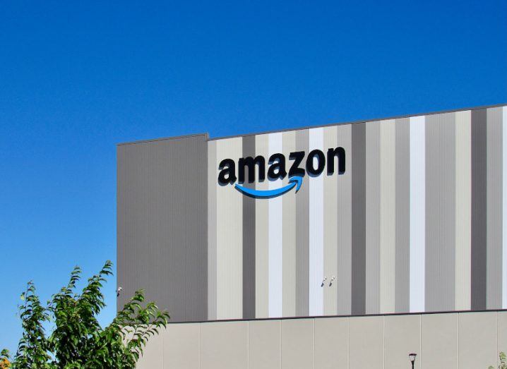 Amazon logo on a warehouse building with blue sky in the background and a tree on the bottom left corner.