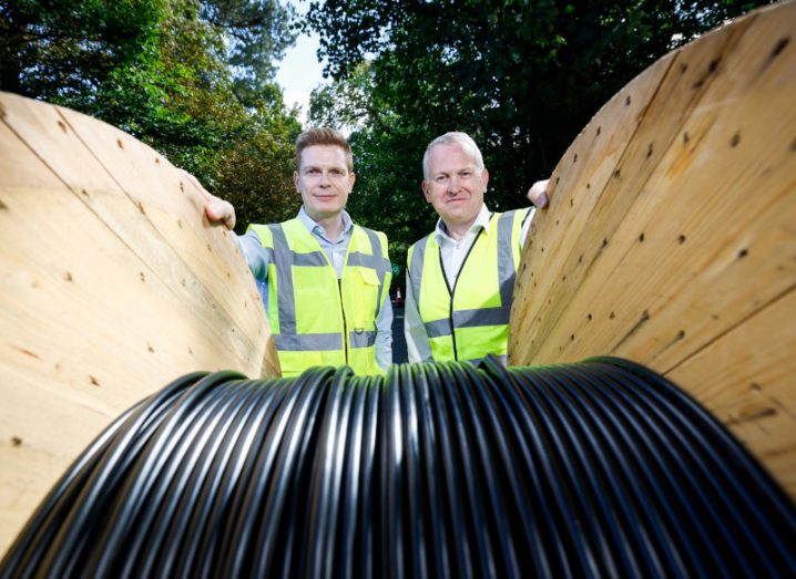 Two men stand behind a big wooden roller with fibre wires wrapped around it.