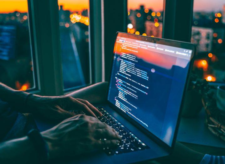 Person working on a laptop with code displayed on the screen. A city skyline at dusk can be seen through glass walls in the background.