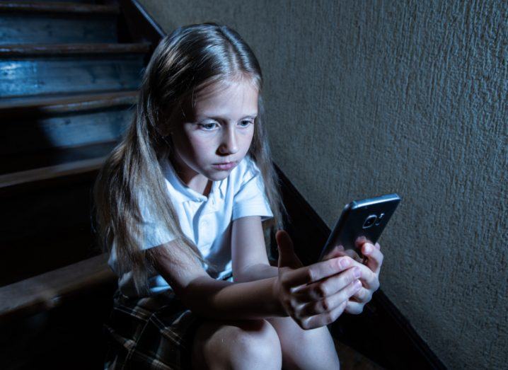 A young sad-looking girl sits on stairs with a smartphone in her hand. Represents cyberbullying.