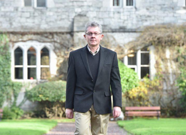 A photo of Professor Séamus Davis walking towards the camera with an ivy-covered university building in the background.