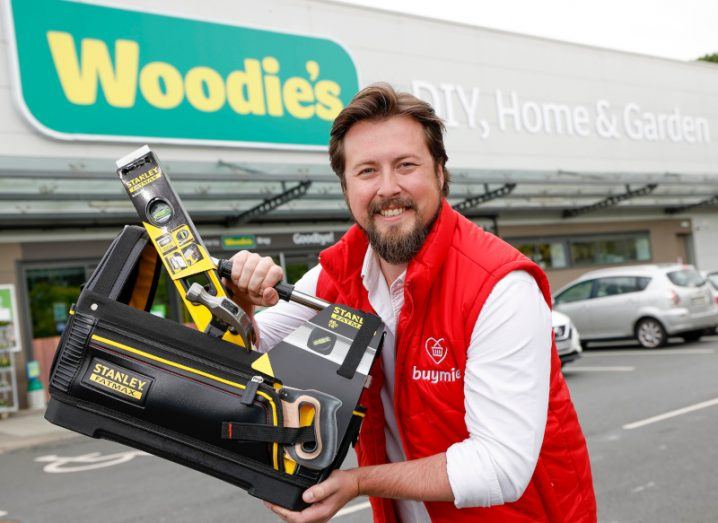 Devan Hughes of Buymie stands outside a Woodie's store holding some DIY equipment.