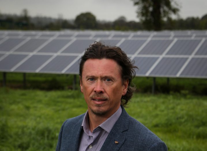 A man in a suit standing in front of some solar panels with grass and trees in the background. He is Strategic Power Projects MD Paul Carson.